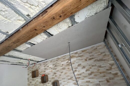 Construction du plafond : isolation apparente, poutres, cloison sèche partielle. Mur de briques texturé avec étagères.